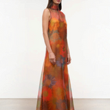 The Albee Dress is an elegant, floor-length gown in sheer orange fabric featuring a colorful, oversized floral print, a sleeveless design, and a high-neck silhouette.