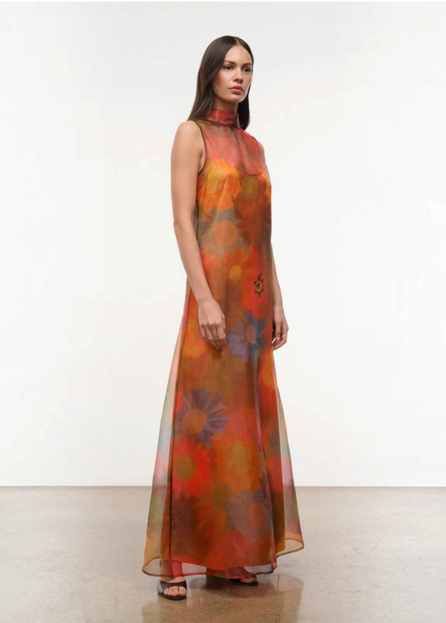 The Albee Dress is an elegant, floor-length gown in sheer orange fabric featuring a colorful, oversized floral print, a sleeveless design, and a high-neck silhouette.