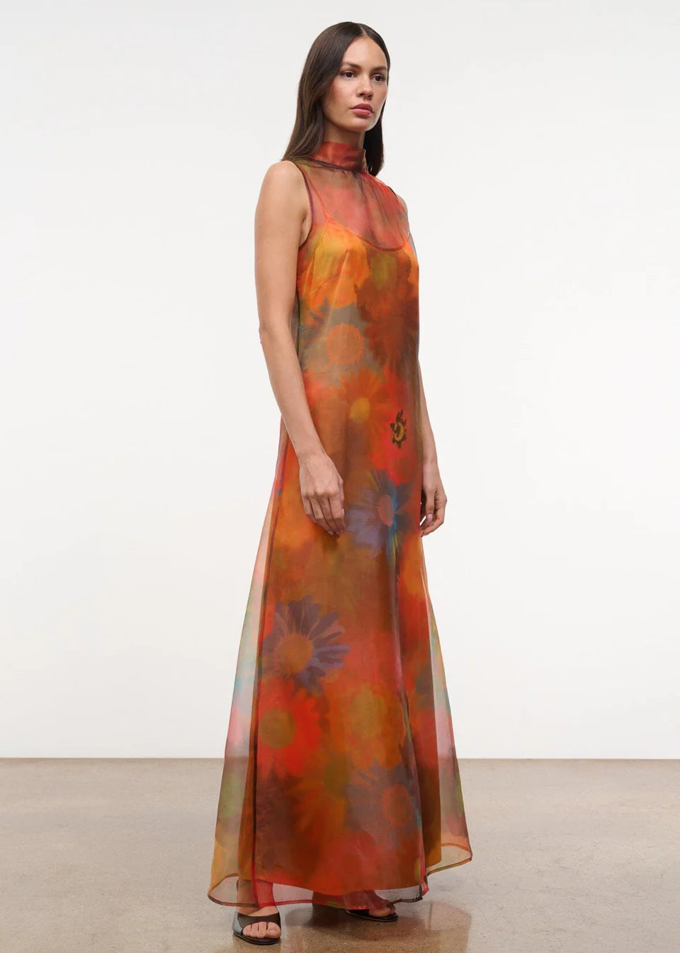 The Albee Dress is an elegant, floor-length gown in sheer orange fabric featuring a colorful, oversized floral print, a sleeveless design, and a high-neck silhouette.