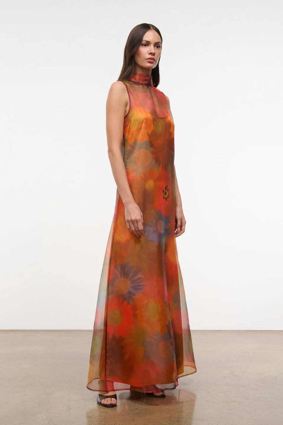 The Albee Dress is an elegant, floor-length gown in sheer orange fabric featuring a colorful, oversized floral print, a sleeveless design, and a high-neck silhouette.