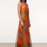 The Albee Dress is an elegant, floor-length gown in sheer orange fabric featuring a colorful, oversized floral print, a sleeveless design, and a high-neck silhouette.