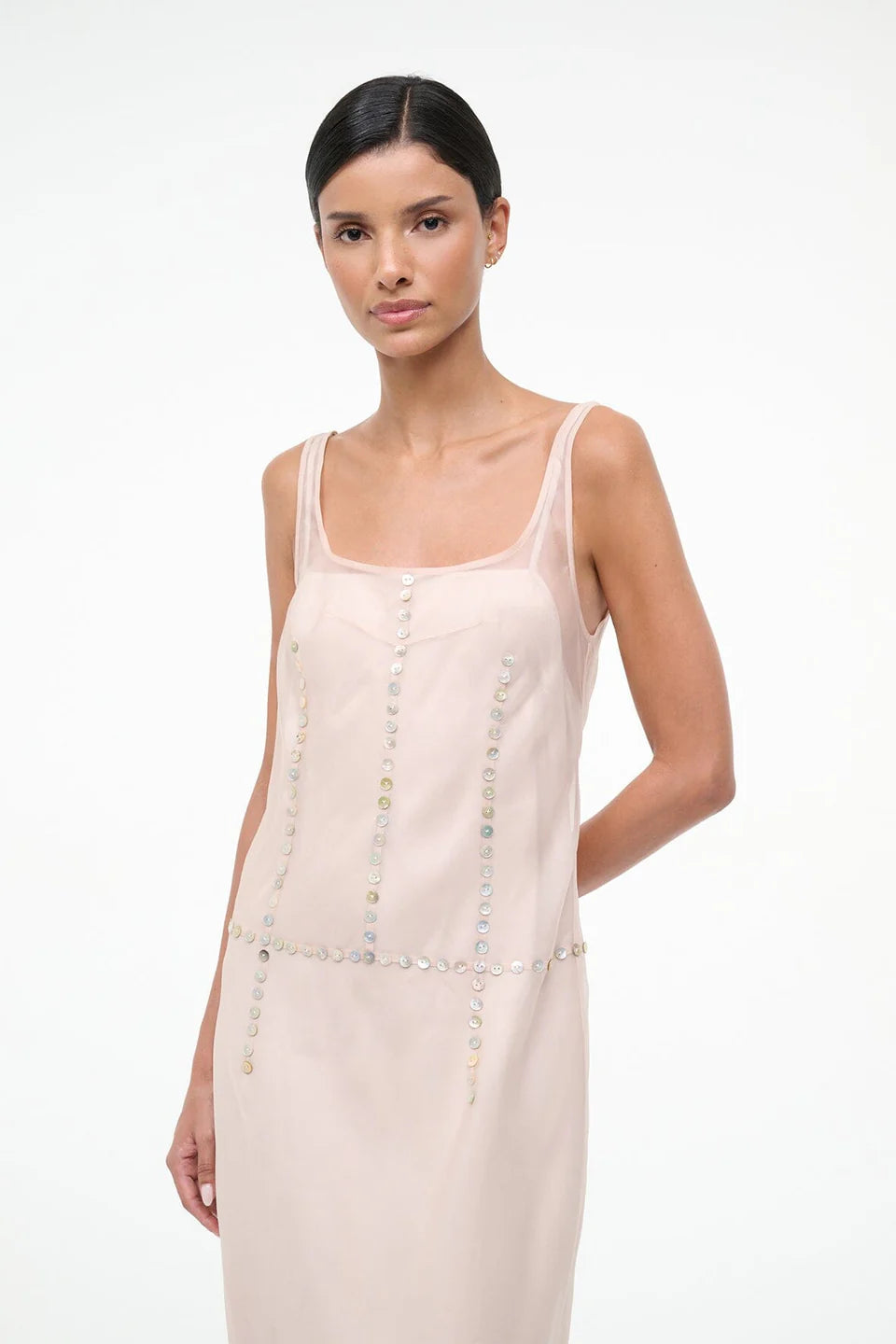 The Greta Dress is an elegant, sleeveless light pink midi dress with a square neckline and rows of small, iridescent buttons, perfect for evening dressing.