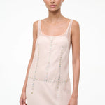 The Greta Dress is an elegant pale pink sleeveless dress with a scoop neckline and vertical and horizontal rows of small, iridescent buttons, perfect for evening dressing.