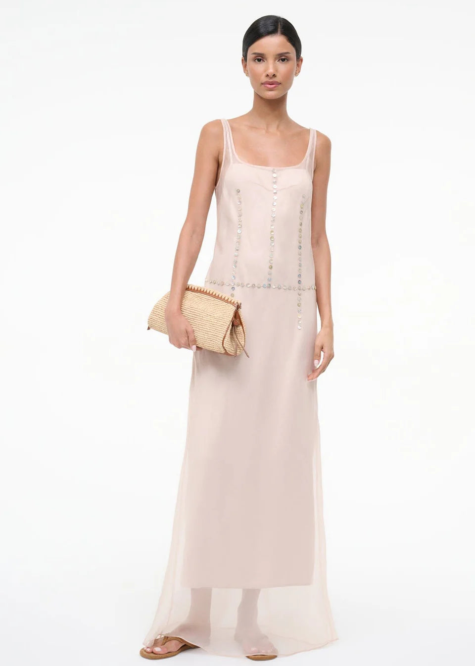 A woman in the pale pink, sheer-layered Greta Dress featuring decorative circular-button trim down the front and waist, holding a woven clutch for special occasions.