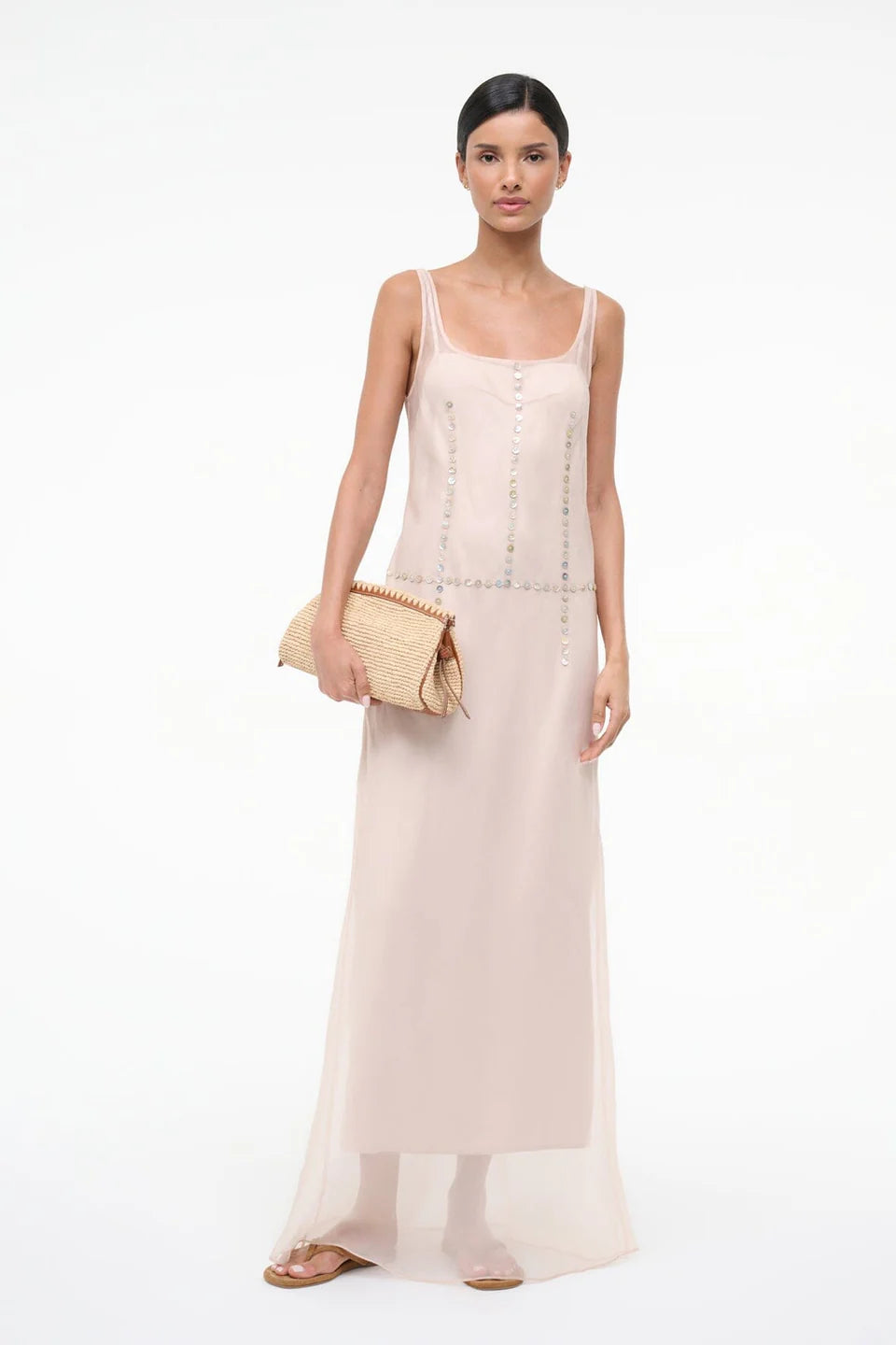 A woman in the pale pink, sheer-layered Greta Dress featuring decorative circular-button trim down the front and waist, holding a woven clutch for special occasions.
