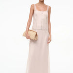 A woman in the pale pink, sheer-layered Greta Dress featuring decorative circular-button trim down the front and waist, holding a woven clutch for special occasions.