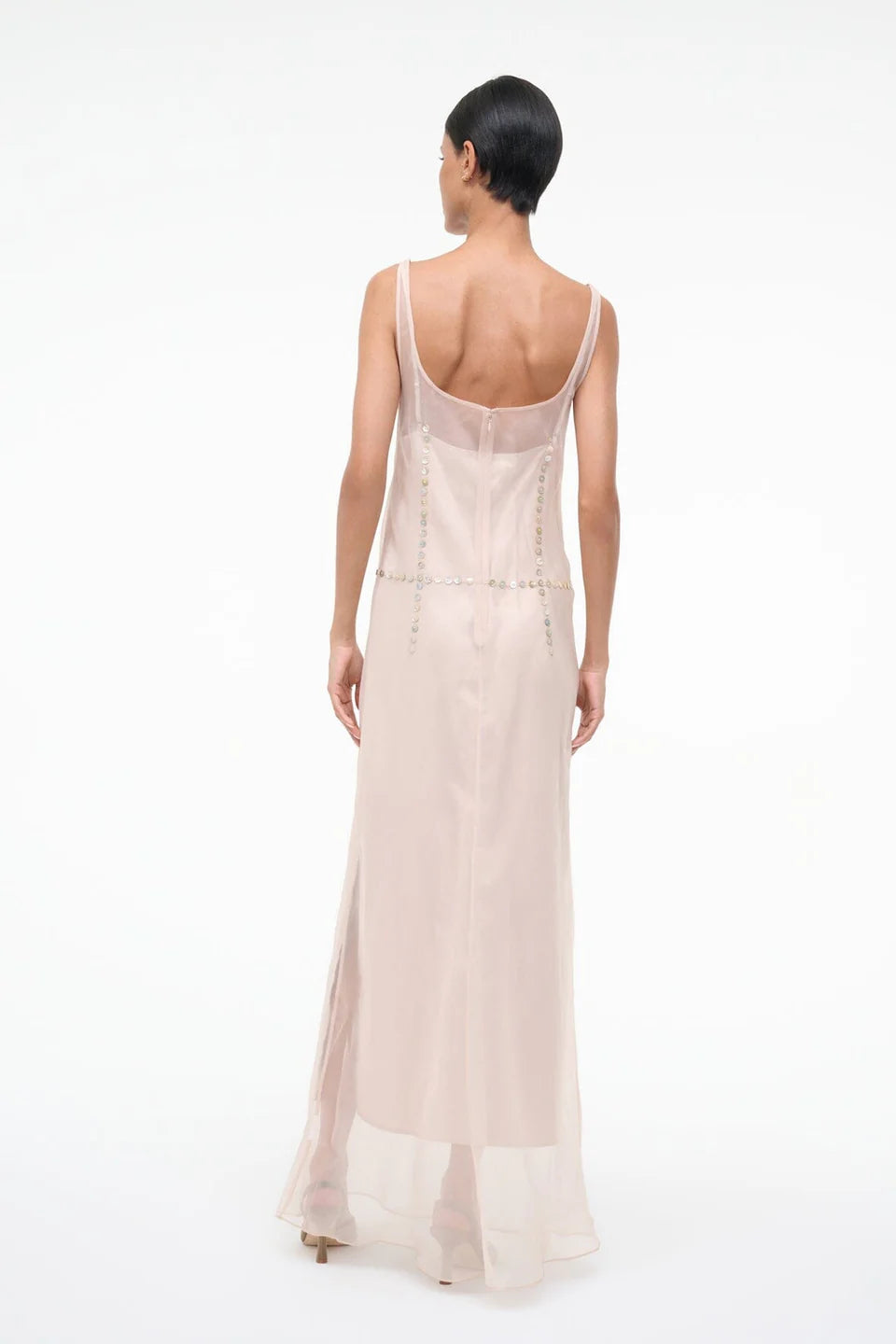 Back view of the Greta Dress, a pale pink sheer maxi dress for special occasions featuring a sleeveless silhouette, scoop back, and metallic disc embellishments at the waist.