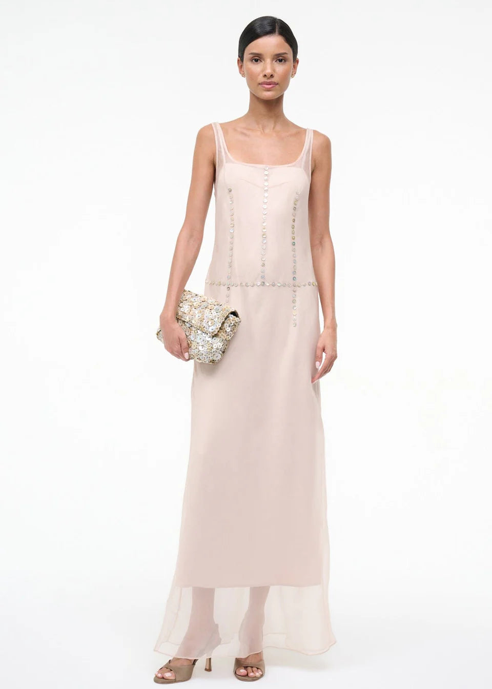 A person wearing the elegant, floor-length light pink Greta Dress, featuring sleeveless styling and circular stud detailing along the seams, while holding a textured clutch bag for special occasions.