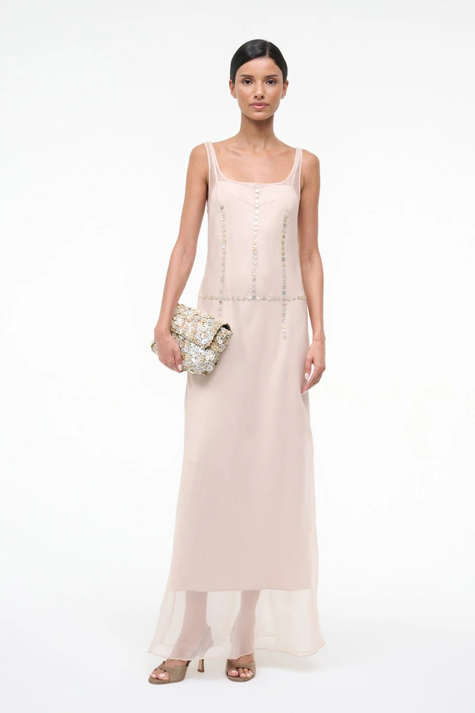 A person wearing the elegant, floor-length light pink Greta Dress, featuring sleeveless styling and circular stud detailing along the seams, while holding a textured clutch bag for special occasions.