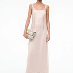 A person wearing the elegant, floor-length light pink Greta Dress, featuring sleeveless styling and circular stud detailing along the seams, while holding a textured clutch bag for special occasions.