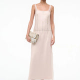 A person wearing the elegant, floor-length light pink Greta Dress, featuring sleeveless styling and circular stud detailing along the seams, while holding a textured clutch bag for special occasions.