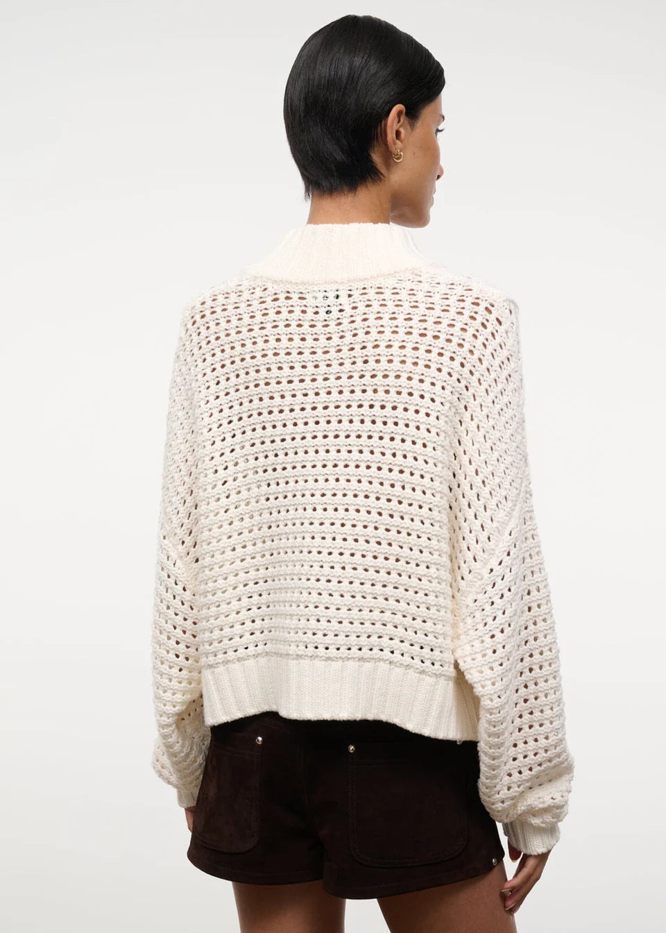 Back view of a person wearing a cream-colored, open-knit Cropped Hampton Sweater featuring a mock neck and ribbed hem.