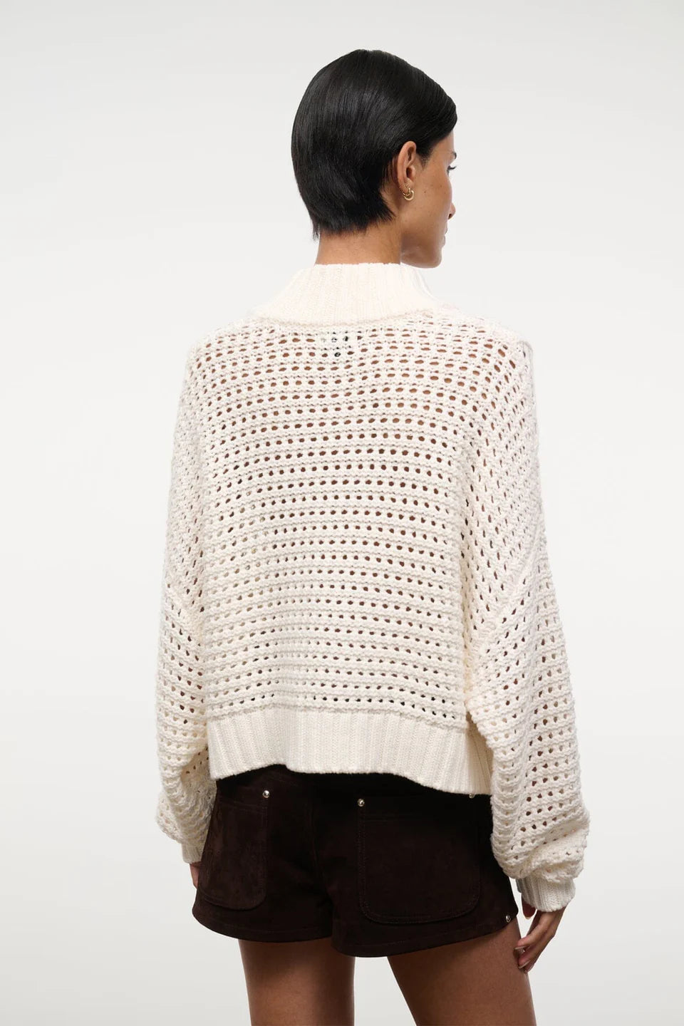 Back view of a person wearing a cream-colored, open-knit Cropped Hampton Sweater featuring a mock neck and ribbed hem.