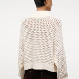 Back view of a person wearing a cream-colored, open-knit Cropped Hampton Sweater featuring a mock neck and ribbed hem.