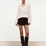 A person wears a versatile cream-colored, long-sleeved, open-knit Cropped Hampton Sweater with a quarter-zip collar, dark brown lace-up shorts, and black knee-high boots.