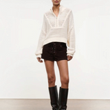A person wears a versatile cream-colored, long-sleeved, open-knit Cropped Hampton Sweater with a quarter-zip collar, dark brown lace-up shorts, and black knee-high boots.