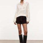A person wears a versatile cream-colored, long-sleeved, open-knit Cropped Hampton Sweater with a quarter-zip collar, dark brown lace-up shorts, and black knee-high boots.