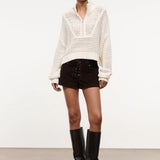 A person wears a versatile cream-colored, long-sleeved, open-knit Cropped Hampton Sweater with a quarter-zip collar, dark brown lace-up shorts, and black knee-high boots.