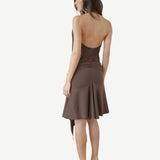 A woman stands facing away, wearing the Halterneck Midi Shell Dress—a brown recycled spandex dress featuring a backless, sleeveless design and flared hem—paired with high heels. Set against a plain white background, this elegant piece is made in London.