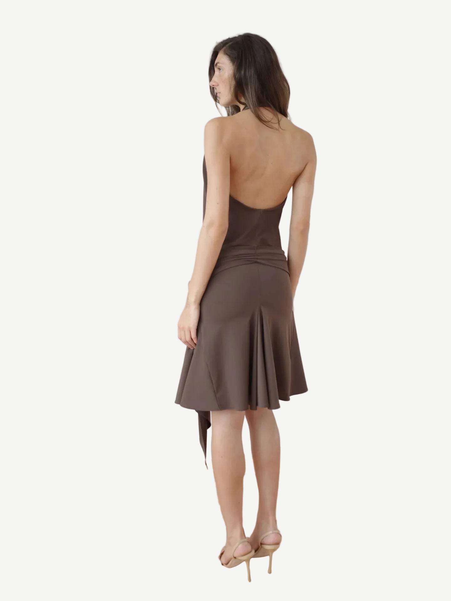 A woman stands facing away, wearing the Halterneck Midi Shell Dress—a brown recycled spandex dress featuring a backless, sleeveless design and flared hem—paired with high heels. Set against a plain white background, this elegant piece is made in London.