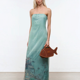 A woman wears the Amara Dress, a strapless, mint green maxi featuring a faint landscape print and subtle sparkles, finished with a brown fish-shaped handbag for an elegant statement.