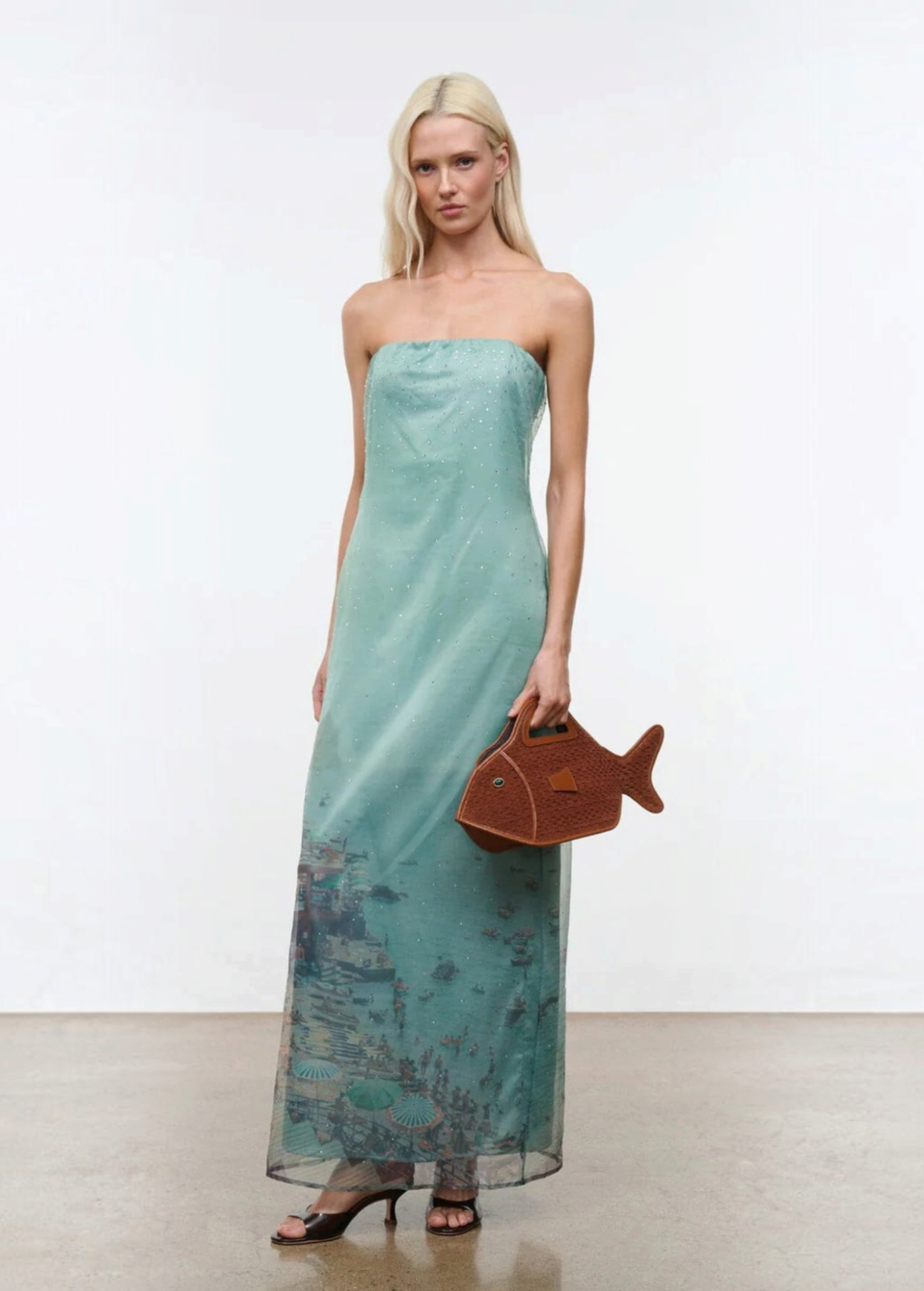 A woman wears the Amara Dress, a strapless, mint green maxi featuring a faint landscape print and subtle sparkles, finished with a brown fish-shaped handbag for an elegant statement.