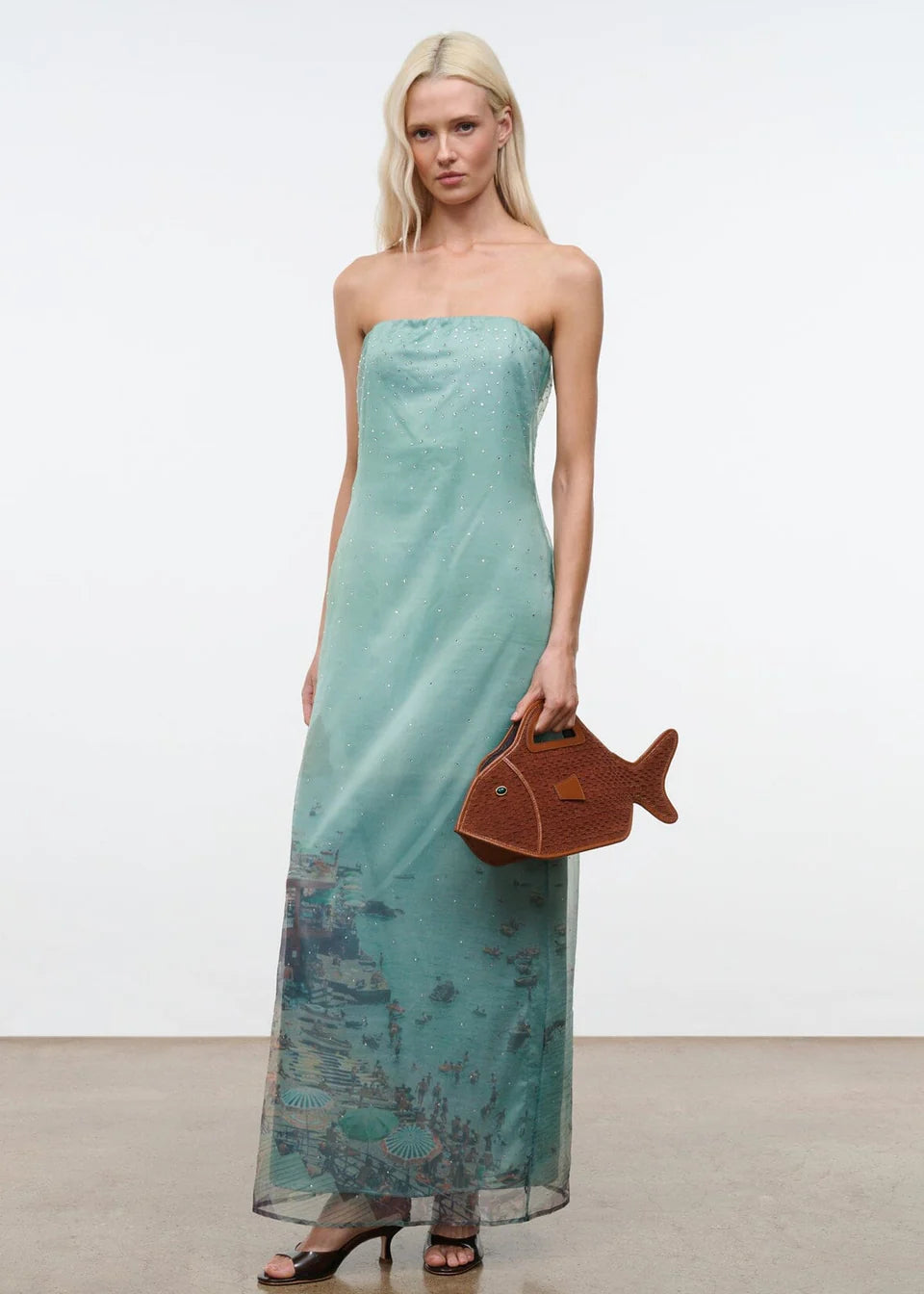 A woman wears the Amara Dress, a strapless, mint green maxi featuring a faint landscape print and subtle sparkles, finished with a brown fish-shaped handbag for an elegant statement.