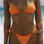 A person wearing the Praia Bottom, an orange triangle bikini bottom made from eco-friendly peach perfect fabric, stands on a sandy beach with ocean waves in the background. Their skin is dusted with sand, and they feature a navel piercing along with two arm bands, showcasing swimwear that effortlessly blends style and sustainability.