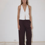 A woman stands against a white wall wearing the Close Halter Top from Deiji Studios, paired with loose dark brown pinstripe pants and black flip-flops. A cable is visible on the floor along the wall.