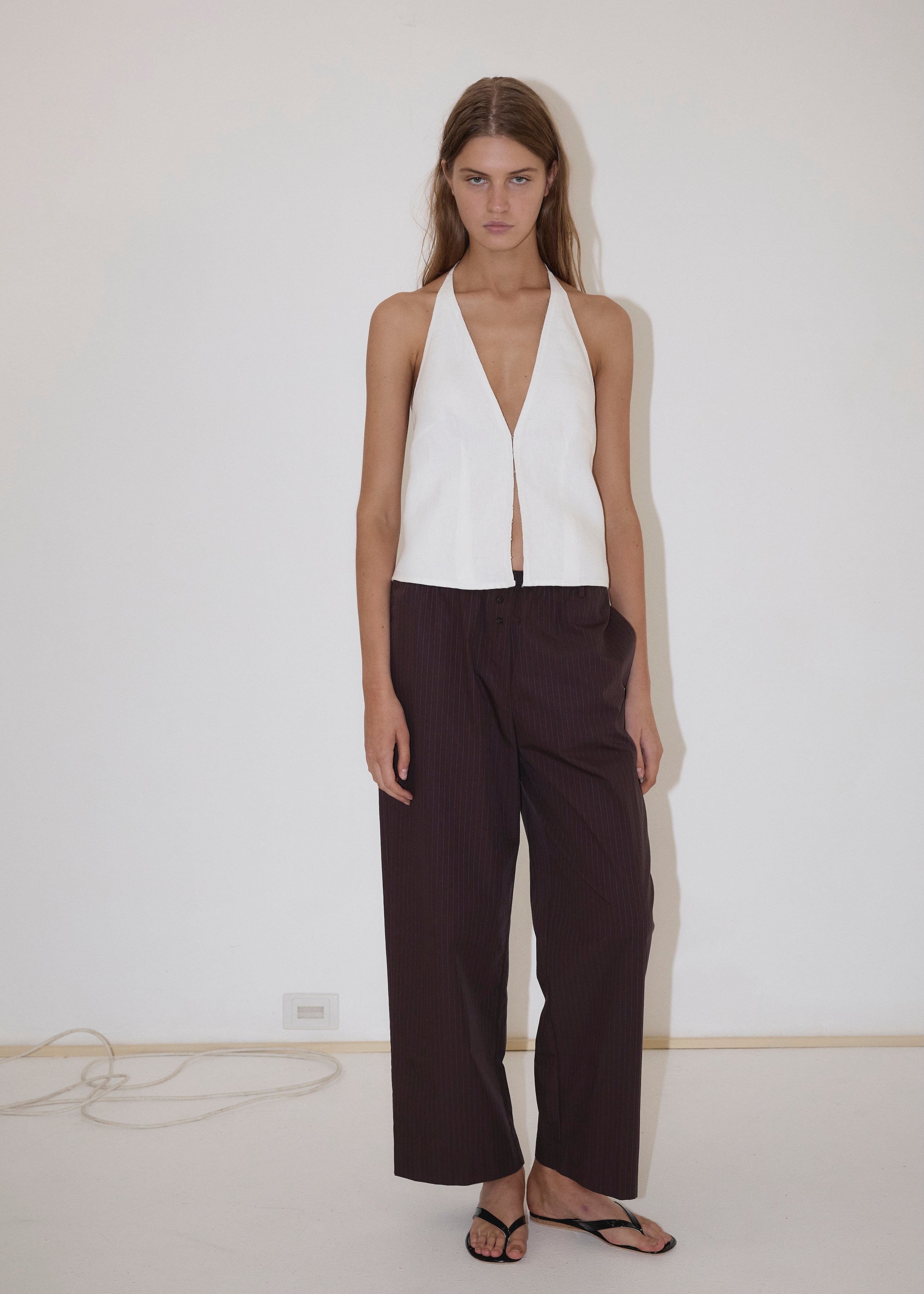 A woman stands against a white wall wearing the Close Halter Top from Deiji Studios, paired with loose dark brown pinstripe pants and black flip-flops. A cable is visible on the floor along the wall.