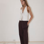 A person stands against a white wall, wearing the Close Halter Top from Deiji Studios—a sleeveless white halter top—paired with loose dark brown pants and black sandals, with hands in their pockets.