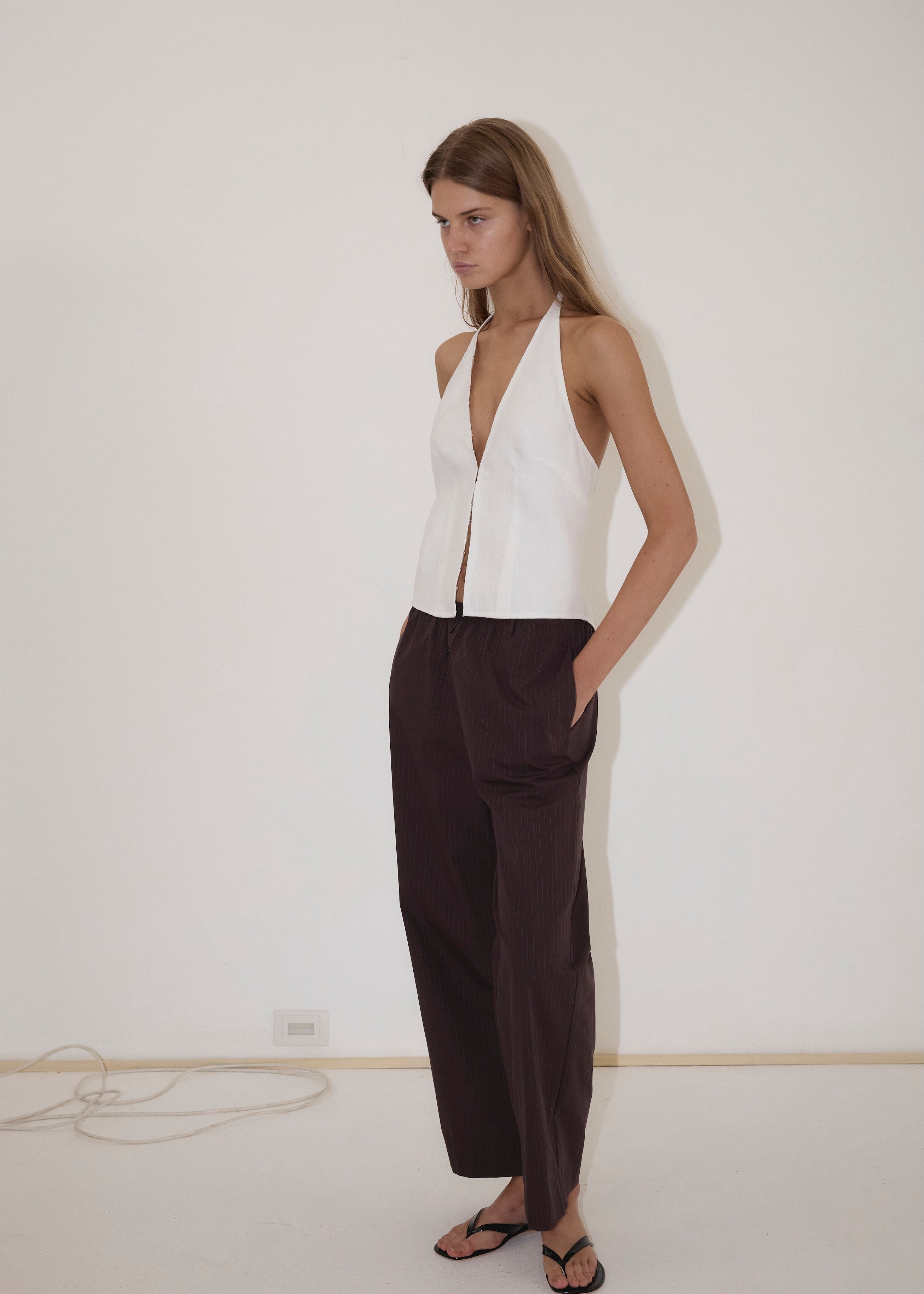 A person stands against a white wall, wearing the Close Halter Top from Deiji Studios—a sleeveless white halter top—paired with loose dark brown pants and black sandals, with hands in their pockets.