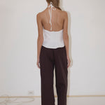 A person stands facing a white wall, wearing the Close Halter Top from Deiji Studios, paired with loose dark pants and black sandals. Their hair is down, and a power cord is visible on the floor near the wall.