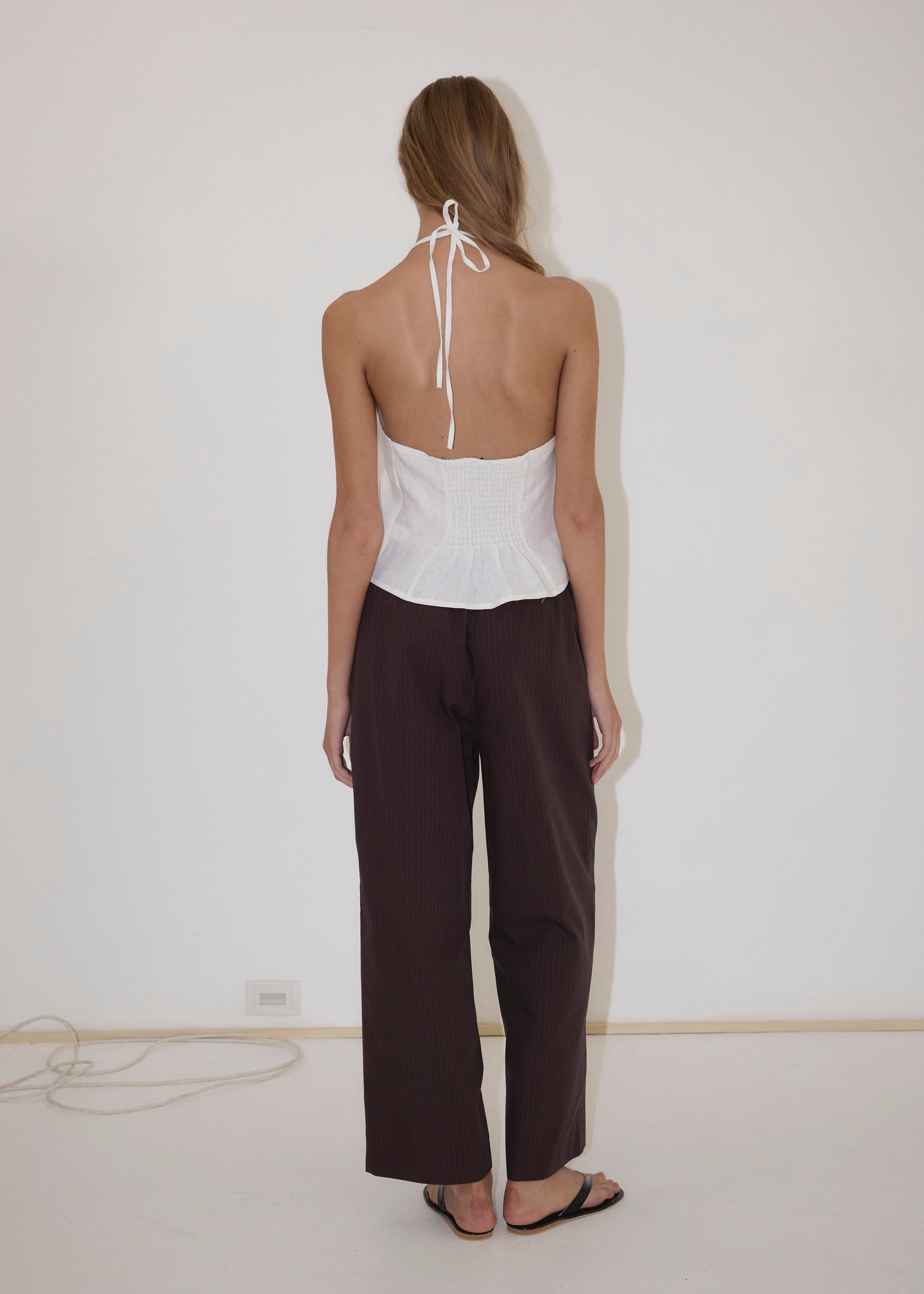 A person stands facing a white wall, wearing the Close Halter Top from Deiji Studios, paired with loose dark pants and black sandals. Their hair is down, and a power cord is visible on the floor near the wall.