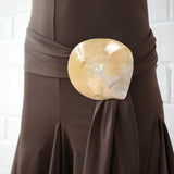 A close-up of the Halterneck Midi Shell Dress, featuring a wide waist sash with a large, shiny seashell buckle at the front. The background is a white brick wall.