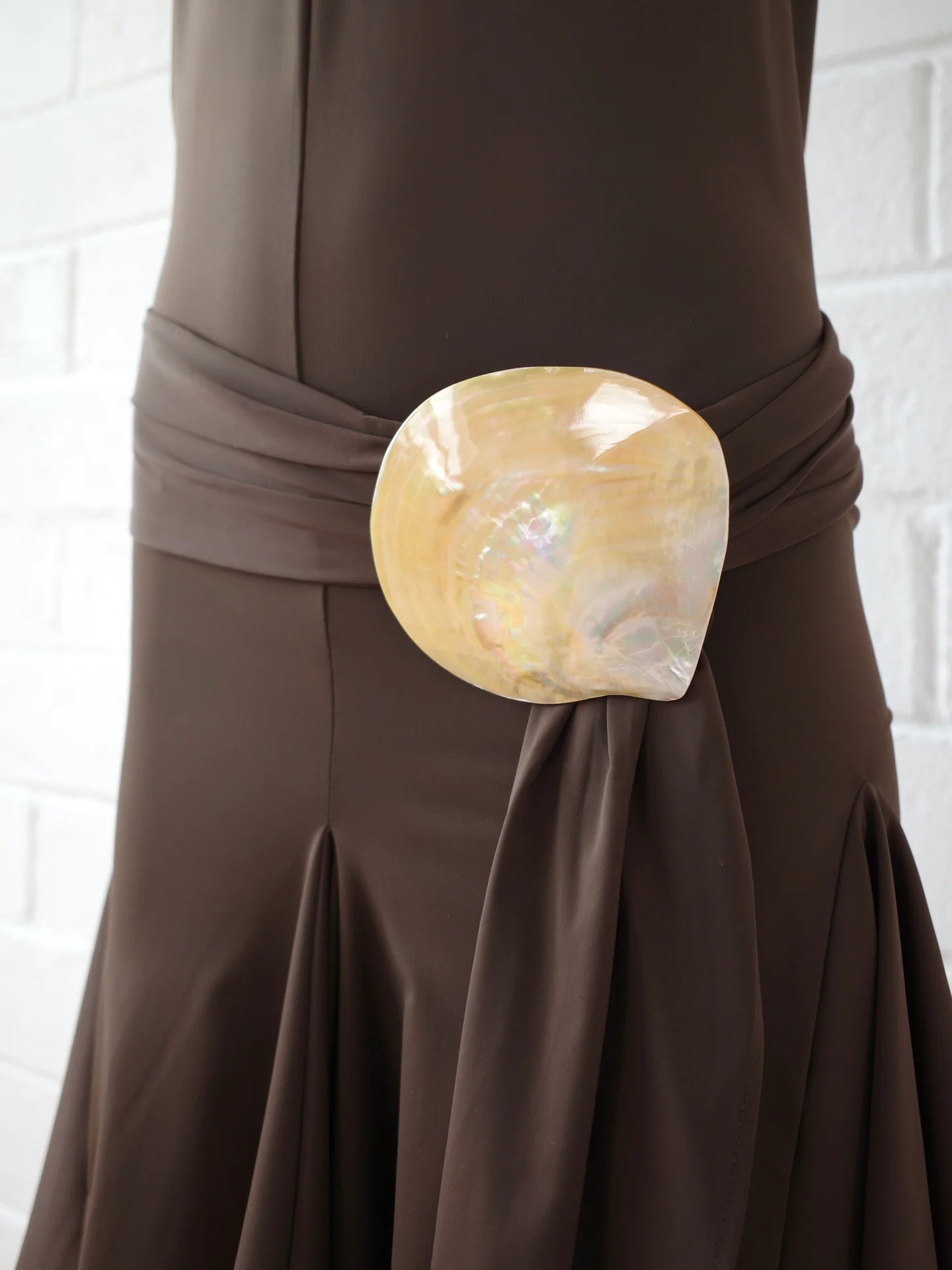 A close-up of the Halterneck Midi Shell Dress, featuring a wide waist sash with a large, shiny seashell buckle at the front. The background is a white brick wall.