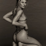 A woman wearing the Praia Bottom, a minimal coverage leopard-print bikini bottom, kneels on the floor, looking over her shoulder at the camera against a plain background. The black and white photo highlights the swimwear’s bold pattern.
