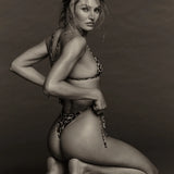 A woman wearing the Praia Bottom, a minimal coverage leopard-print bikini bottom, kneels on the floor, looking over her shoulder at the camera against a plain background. The black and white photo highlights the swimwear’s bold pattern.