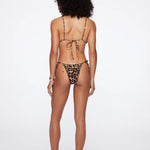 A woman stands with her back to the camera on a plain white background, wearing the Praia Bottom bikini in leopard print with adjustable side ties, paired with black slides.