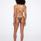 A woman stands with her back to the camera on a plain white background, wearing the Praia Bottom bikini in leopard print with adjustable side ties, paired with black slides.