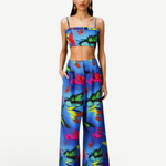 A woman models the Alma Set—a blue, tropical floral print linen ensemble featuring a crop top and matching wide-leg pants—standing with hands in her pockets against a white background, highlighting its versatile styling potential with adaptable accessories.