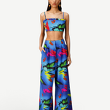 A woman models the Alma Set—a blue, tropical floral print linen ensemble featuring a crop top and matching wide-leg pants—standing with hands in her pockets against a white background, highlighting its versatile styling potential with adaptable accessories.