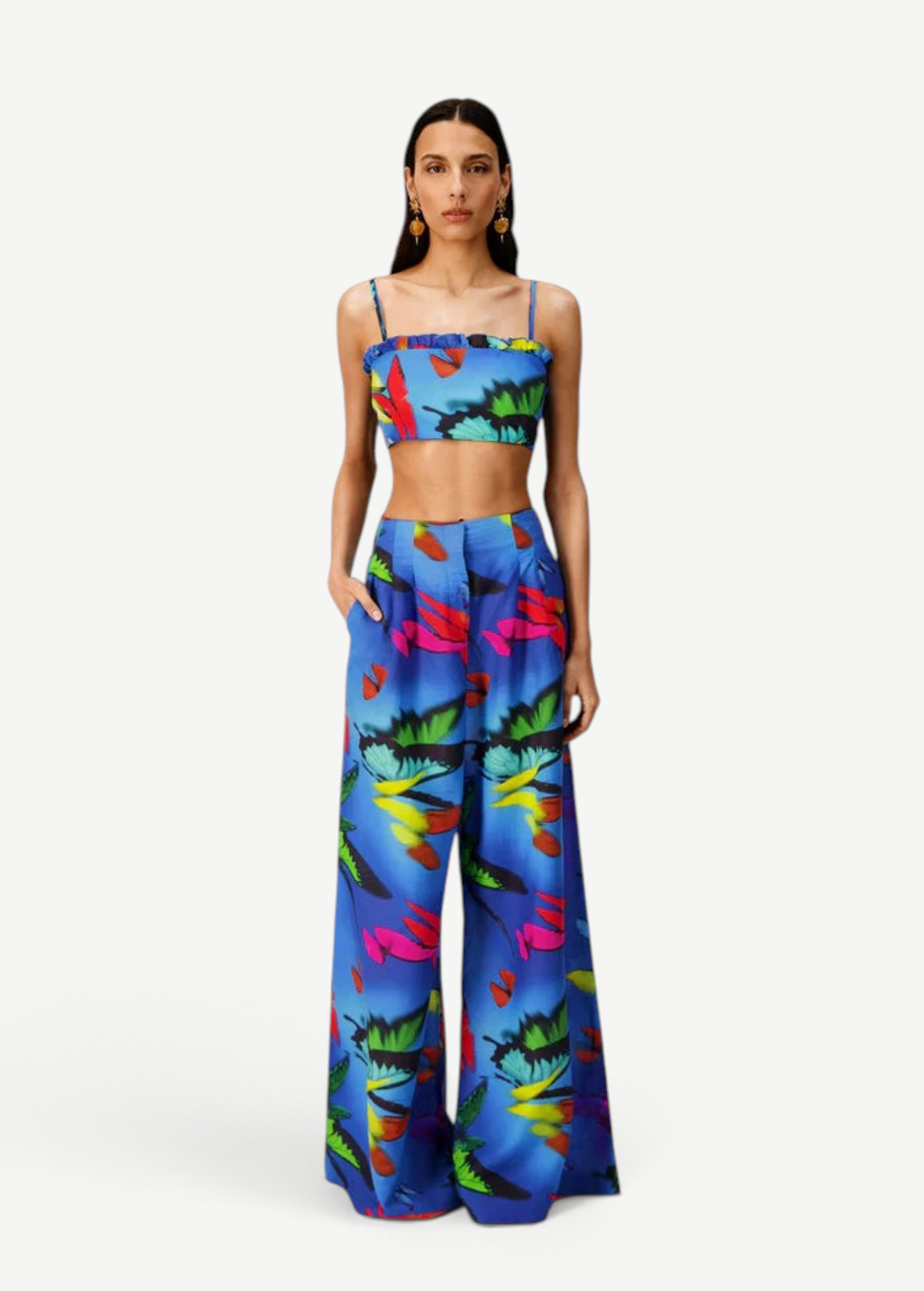 A woman models the Alma Set—a blue, tropical floral print linen ensemble featuring a crop top and matching wide-leg pants—standing with hands in her pockets against a white background, highlighting its versatile styling potential with adaptable accessories.
