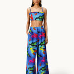 A woman models the Alma Set—a blue, tropical floral print linen ensemble featuring a crop top and matching wide-leg pants—standing with hands in her pockets against a white background, highlighting its versatile styling potential with adaptable accessories.