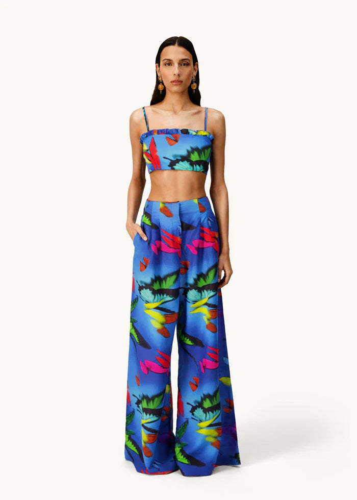 A woman models the Alma Set—a blue, tropical floral print linen ensemble featuring a crop top and matching wide-leg pants—standing with hands in her pockets against a white background, highlighting its versatile styling potential with adaptable accessories.