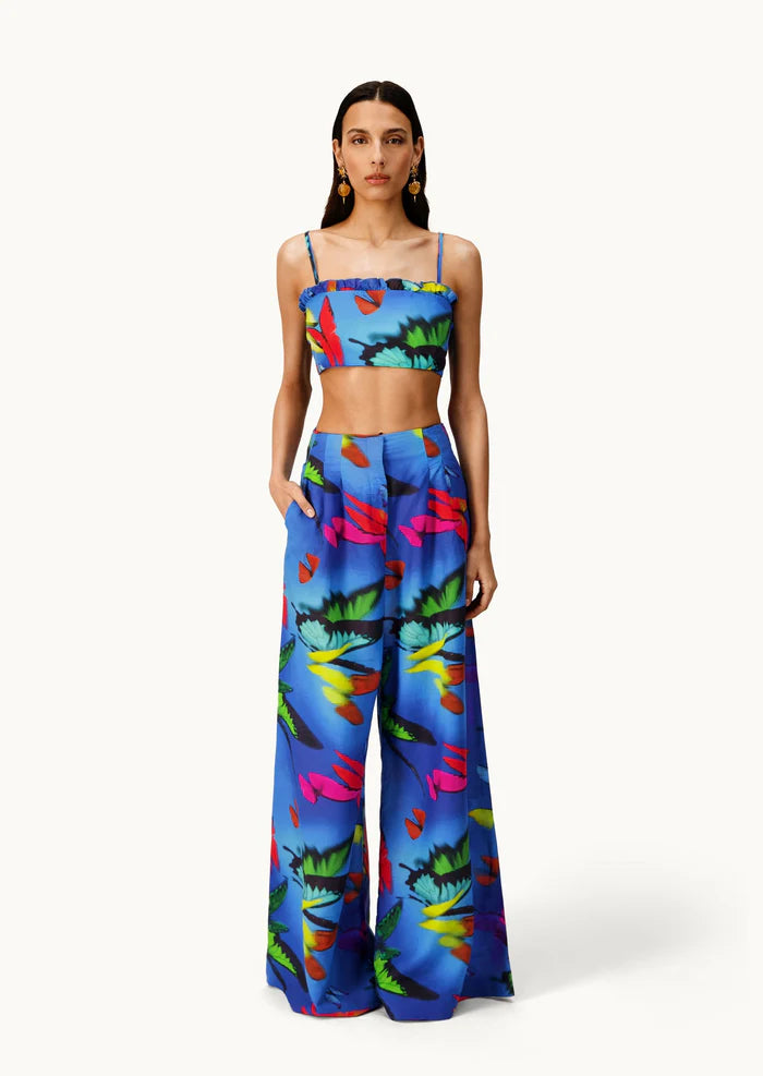 A woman models the Alma Set—a blue, tropical floral print linen ensemble featuring a crop top and matching wide-leg pants—standing with hands in her pockets against a white background, highlighting its versatile styling potential with adaptable accessories.