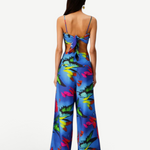 A woman faces away, wearing the Alma Set—a blue floral jumpsuit with wide-leg pants and an open back finished with a statement bow. Her long, straight hair pairs with a single hoop earring, showcasing the set’s versatile styling that can be effortlessly elevated for any occasion with adaptable accessories.