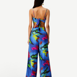 A woman faces away, wearing the Alma Set—a blue floral jumpsuit with wide-leg pants and an open back finished with a statement bow. Her long, straight hair pairs with a single hoop earring, showcasing the set’s versatile styling that can be effortlessly elevated for any occasion with adaptable accessories.