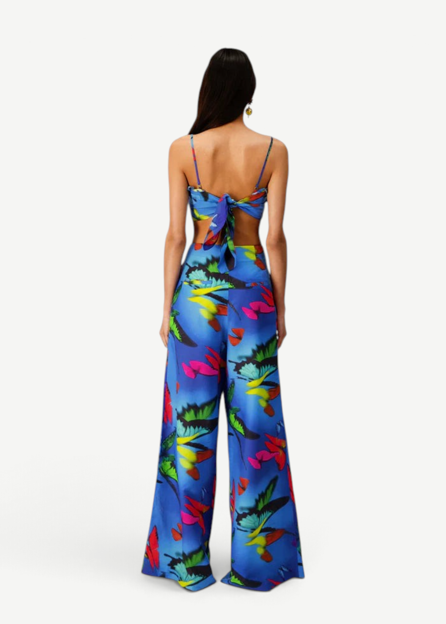 A woman faces away, wearing the Alma Set—a blue floral jumpsuit with wide-leg pants and an open back finished with a statement bow. Her long, straight hair pairs with a single hoop earring, showcasing the set’s versatile styling that can be effortlessly elevated for any occasion with adaptable accessories.