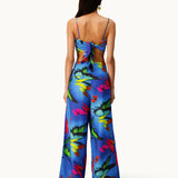 A woman faces away, wearing the Alma Set—a blue floral jumpsuit with wide-leg pants and an open back finished with a statement bow. Her long, straight hair pairs with a single hoop earring, showcasing the set’s versatile styling that can be effortlessly elevated for any occasion with adaptable accessories.